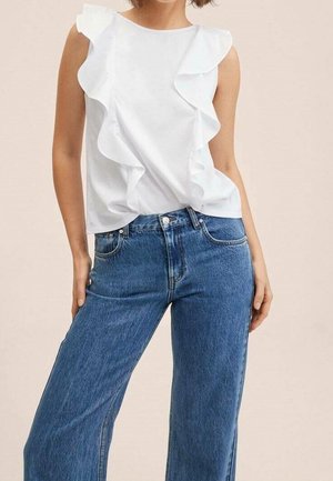 Femme portant un haut blanc sans manches avec un devant à volants et un jean en denim bleu taille haute, debout avec une main dans la poche.