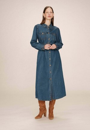 Robe chemise en jean à manches longues, col, poches poitrine et boutonnage à l’avant. Tissu bleu foncé, longueur genoux, associée à des bottes à talons marron.