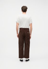 Άνδρας με την πλάτη στραμμένη, φοράει λευκό μπλουζάκι, καφέ παντελόνια κορδούρας ευρείας γραμμής με το κείμενο «HOUSE OF SUNNY» στην πίσω τσέπη, μαύρα αθλητικά παπούτσια και λευκές κάλτσες.