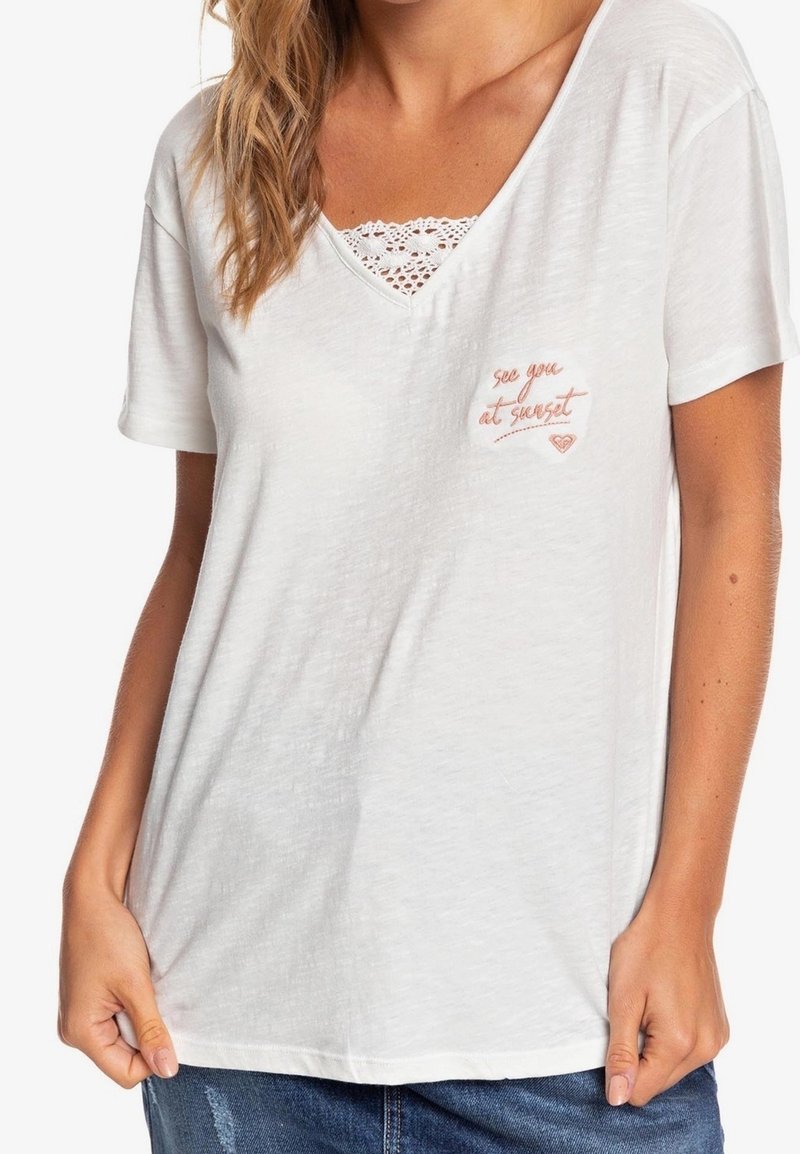 Femme portant un T-shirt blanc à col V avec texte brodé « see you at sunset » et détail en dentelle en dessous, associé à un jean denim bleu.