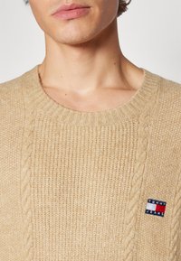 Pull en tricot beige avec un col rond côtelé et un motif en cable. Présente un petit patch logo Tommy Jeans. Texture douce et coutures détaillées.