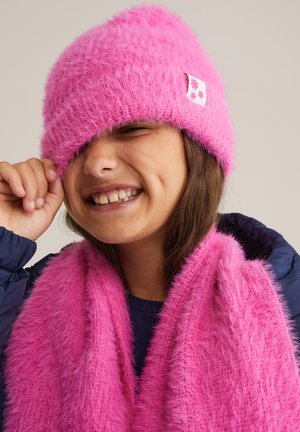 Rozā pūkains adīts cepure, kas sedz acis, ar kvadrātveida etiķeti ar ziedu dizainu; atbilstoša rozā šalle ar mīkstu tekstūru, kas ietītas ap kaklu.