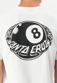 Person som har på seg en hvit t-skjorte med stor svart og hvit 8-ball grafikk og "Santa Cruz" tekst på baksiden.