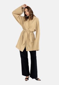 Beżowa, oversize'owa kurtka z szerokimi rękawami i zawiązywanym w pasie paskiem, zestawiona z czarnymi spodniami o szerokim kroju i czarnymi sandałami na wysokim obcasie.