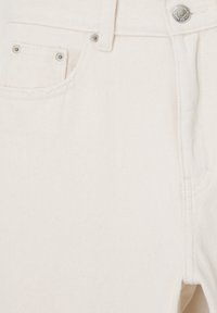 Gros plan sur un jean en denim blanc cassé montrant une poche avant, des rivets en métal, une boucle de ceinture et une fermeture par bouton métallique.