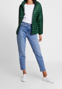 Grøn quiltet jakke med høj krave, hvid t-shirt under, blå højttaljede jeans og hvide sneakers. Glat tekstur, afslappet pasform.