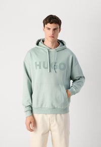 Giovane uomo che indossa una felpa verde chiaro HUGO con tasca frontale e pantaloni crema, in piedi contro uno sfondo chiaro e uniforme.