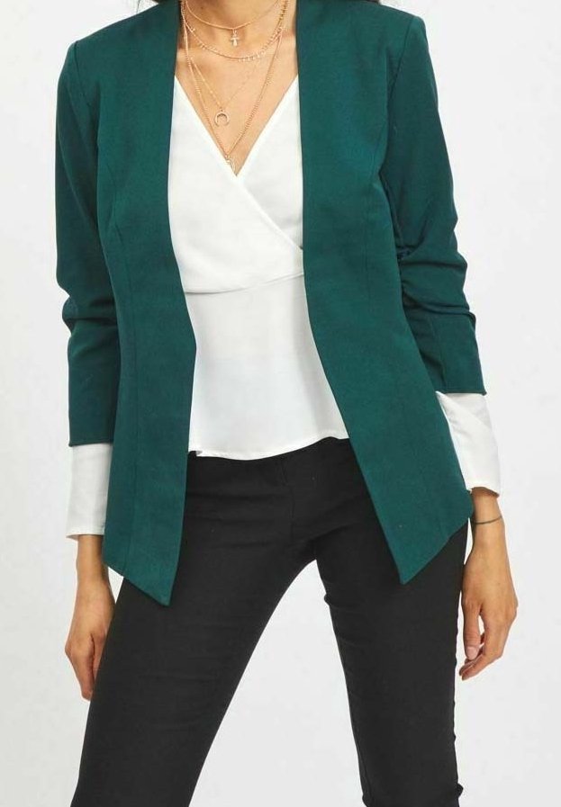 Femme portant un blazer ouvert vert foncé, un chemisier blanc portefeuille, un pantalon noir et des colliers dorés superposés avec des pendentifs en forme de croix et de croissant.