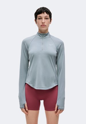 Jeune personne aux cheveux courts et foncés portant un haut zippé bleu clair à manches longues et un short moulant rouge foncé, debout devant un fond uni.