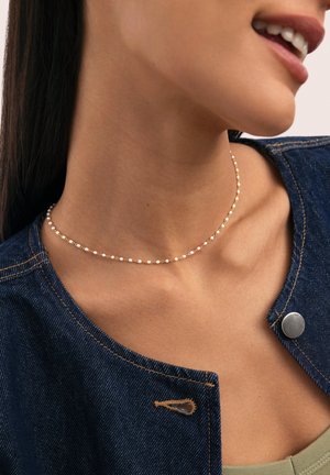 Mujer con un delicado collar de cadena dorada con pequeñas cuentas blancas, combinado con una chaqueta vaquera azul oscuro y una camiseta verde.