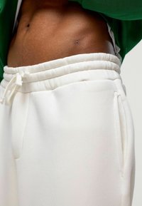 Witte joggingbroek met een elastische tailleband, trekkoordsluiting en zijzakken, gepresenteerd tegen een huidkleurige top en groene top.