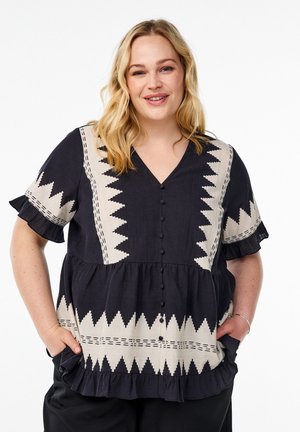 Blusa corta de manga corta en negro y crema con escote en V, que presenta un patrón en zigzag y bordes con volantes. Cierre de botones en el frontal y bolsillos laterales.