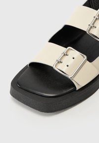 Svarte og kremfargede sandaler med en bred kremfarget stropp som har en sølvspenne; teksturert innersåle og tykk svart gummisåle.