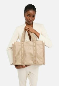 Beige fabric tote bag with plush texture, gold studs, and embossed "LIU JO" logo. Features sturdy handles for easy carry.
