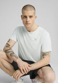 Lyseblå atletisk T-shirt med rund halsudskæring, korte ærmer og et lille gråt logo, parret med sorte shorts i en siddende position.