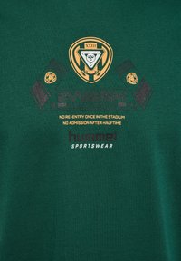 Grüner Stoff mit goldenem und schwarzem Sportlogo, Schild, Schals und Text mit Stadion-Wiedereintrittsregeln sowie "hummel SPORTSWEAR"-Markenbranding.