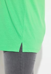Groene katoenen t-shirt met een splitzoom, gecombineerd met grijze slim fit broek. De stof is soepel en het ontwerp is casual en losvallend.