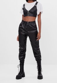 Kvinna i svart lädercroppad zip-up topp, matchande högmidjade joggerbyxor och svarta snörade platåsneakers mot vit bakgrund.