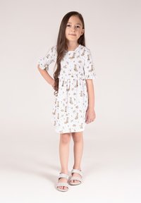 Biała sukienka z krótkimi rękawami, z nadrukiem brązowych zwierząt i kwiatów, zebrana w talii, z falbankami na rękawach. Noszona ze srebrnymi sandałami.