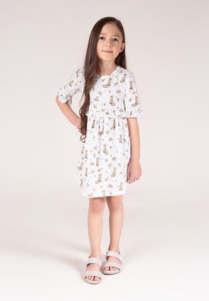 Biała sukienka z krótkimi rękawami, z nadrukiem brązowych zwierząt i kwiatów, zebrana w talii, z falbankami na rękawach. Noszona ze srebrnymi sandałami.