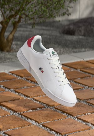 Baskets blanches Lacoste avec languette de talon rouge et logo crocodile vert, posées sur des carreaux carrés en bois et de petits cailloux gris en extérieur.