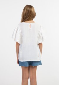 Weiße Baumwollbluse mit Flatterärmeln, einem Schlüssellochverschluss am Rücken und gestickten Details an den Seiten, kombiniert mit Jeansshorts.