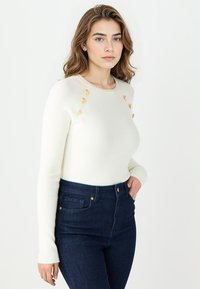 Pull en tricot blanc avec texture côtelée, décolleté rond et accents de boutons dorés sur les épaules. Associé à un jean bleu foncé taille haute.