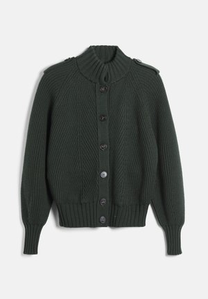 Femme portant un cardigan tricoté côtelé vert foncé avec de gros boutons et un pantalon vert assorti, debout sur un fond uni.