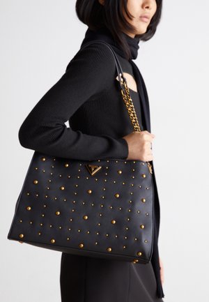 Femme portant un haut noir à manches longues porte un sac à main en cuir noir avec des clous dorés et une bandoulière chaîne sur l'épaule.