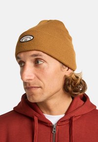 Bruine ribgebreide beanie met een logopatch aan de voorkant. Gekozen bij een rode zip-up hoodie, met daaronder een witte T-shirt. Zachte gebreide textuur.