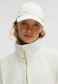 Ženska z svetlo kodrasto lasmi nosi belo jakno na zadrgo in belo kapo z diskretno "lululemon" oznako na enobarvni podlagi.