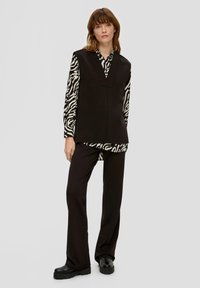 Schwarze ärmellose Tunika, die über einem langärmeligen, schwarz-weißen Zebramuster-Shirt getragen wird; kombiniert mit schwarzen geraden Hosen und klobigen Schuhen.