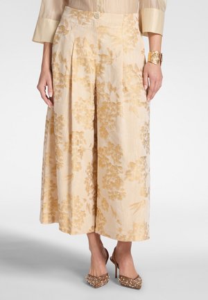 Pantalones beige de pierna ancha y corte cropped con estampado floral dorado, combinados con top beige transparente, pulsera rígida dorada y tacones puntiagudos adornados.