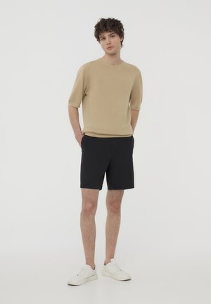 Maglione a maniche corte beige in maglia abbinato a pantaloni neri. Il modello indossa sneakers bianche. Design minimalista e schema di colori solidi.