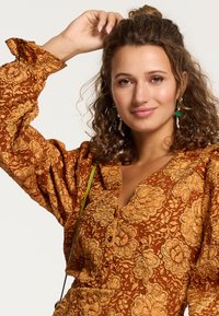 Braunes blumenmusterndes Kleid mit goldenen Akzenten, Puffärmeln, V-Ausschnitt und einem strukturierten Stoff. Grüne Ohrringe und ein Crossbody-Tragegurt sind sichtbar.