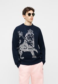 Joven vestido con suéter azul oscuro con un gran emblema de león claro y pantalones rosa claro, de pie con las manos en los bolsillos.