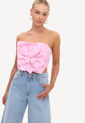 Femme debout portant un haut rose sans bretelles avec un large détail fleur en tissu et un jean bleu clair taille haute à jambes larges sur fond blanc.