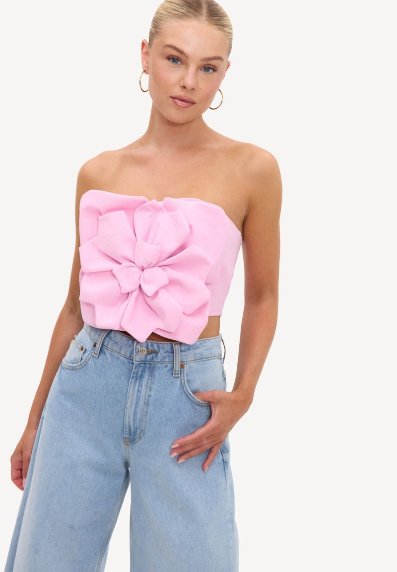 Femme debout portant un haut rose sans bretelles avec un large détail fleur en tissu et un jean bleu clair taille haute à jambes larges sur fond blanc.