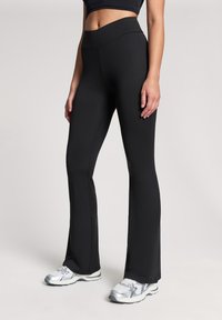 Czarne legginsy z wysokim stanem i rozszerzanymi nogawkami, wykonane z elastycznego materiału, charakteryzujące się gładką teksturą i dopasowanym krojem, w połączeniu z srebrnymi butami sportowymi.