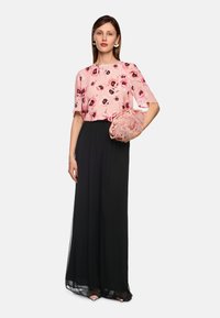 Blumige rosa Bluse mit kurzen Ärmeln, kombiniert mit einem hoch taillierten schwarzen Rock. Das Model hält eine blushfarbenen, federbesetzten Clutch.