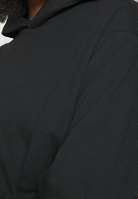 Personne portant un sweat à capuche noir avec une capuche et une épaule visibles, cheveux bouclés légèrement visibles au col.