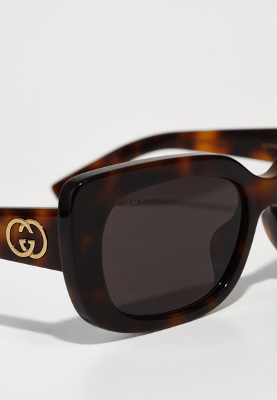 Lunettes de soleil en écaille brune avec des verres carrés, arborant un logo Gucci doré sur le côté et des verres foncés. Texture lisse sur l'ensemble.