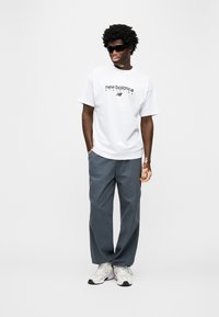 Λευκό μπλουζάκι με το λογότυπο "new balance", γκρι παντελόνι φαρδιάς γραμμής και λευκά αθλητικά παπούτσια με γκρι λεπτομέρειες. Το μοντέλο φοράει μαύρα γυαλιά ηλίου και στέκεται σε στάση.