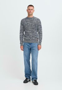 Vyras stovi atsisukęs į priekį, vilkėdamas mėlynai ir baltai dėmėtą megztinį, šviesiai mėlynas džinsus ir juodus sportbačius su baltais dryžiais.