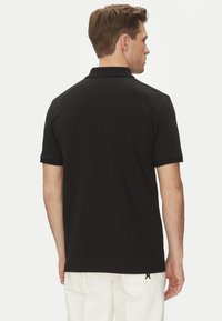 Polo shirt nero a maniche corte con colletto a coste, design posteriore semplice e dettagli di cucitura sottili. Indossato con pantaloni bianchi.