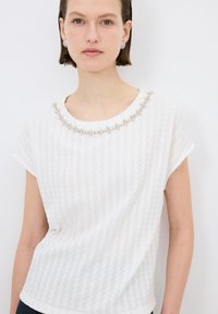 Top blanco de manga corta con un patrón texturizado y un cuello decorativo con cuentas. La tela es ligera y suave.