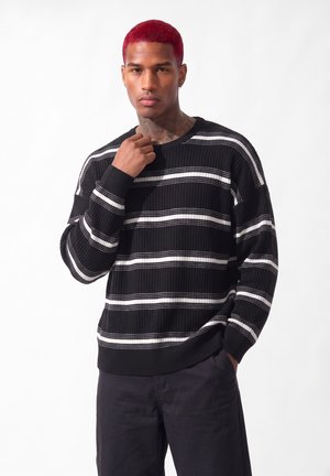 Homme aux cheveux rouges portant un pull rayé noir et blanc et un pantalon noir, debout devant un fond blanc uni.