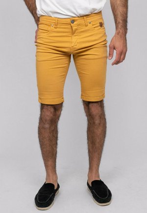 Homme portant un short jaune moutarde longueur genou, une chemise blanche et des chaussures noires sans lacets, debout devant un fond uni.