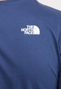 Mörkblå t-shirt med kort ärm i bomull; har en vit logotyp av The North Face på övre vänstra bröstområdet. Slät textur.