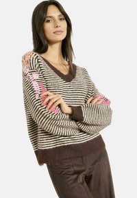 Maglione a righe di maglia in beige e marrone con scollatura a V profonda e rifiniture marroni. Presenta un ricamo floreale colorato sulla manica sinistra.